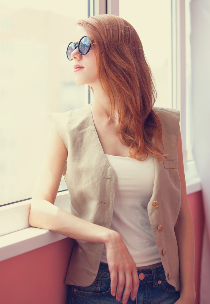 Beautiful redheaded lady looking out a window while wearing a pair of fashionable sunglasses